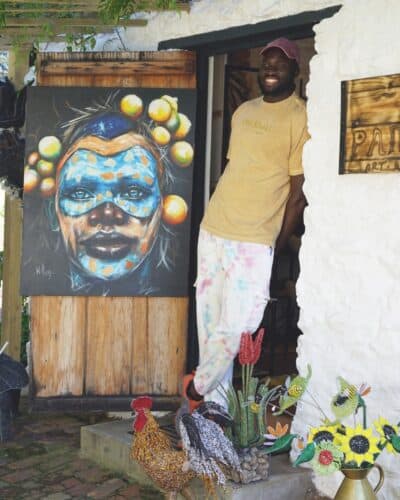 Paivepo's founder standing at the door of his store next to an artwork at Old Nick Village
